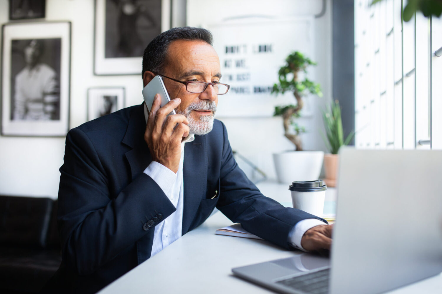 Serious concentrated busy old caucasian man in suit, glasses calls by phone, typing on laptop in office interior. Multitasking, modern work, business remotely, app for meeting, communication